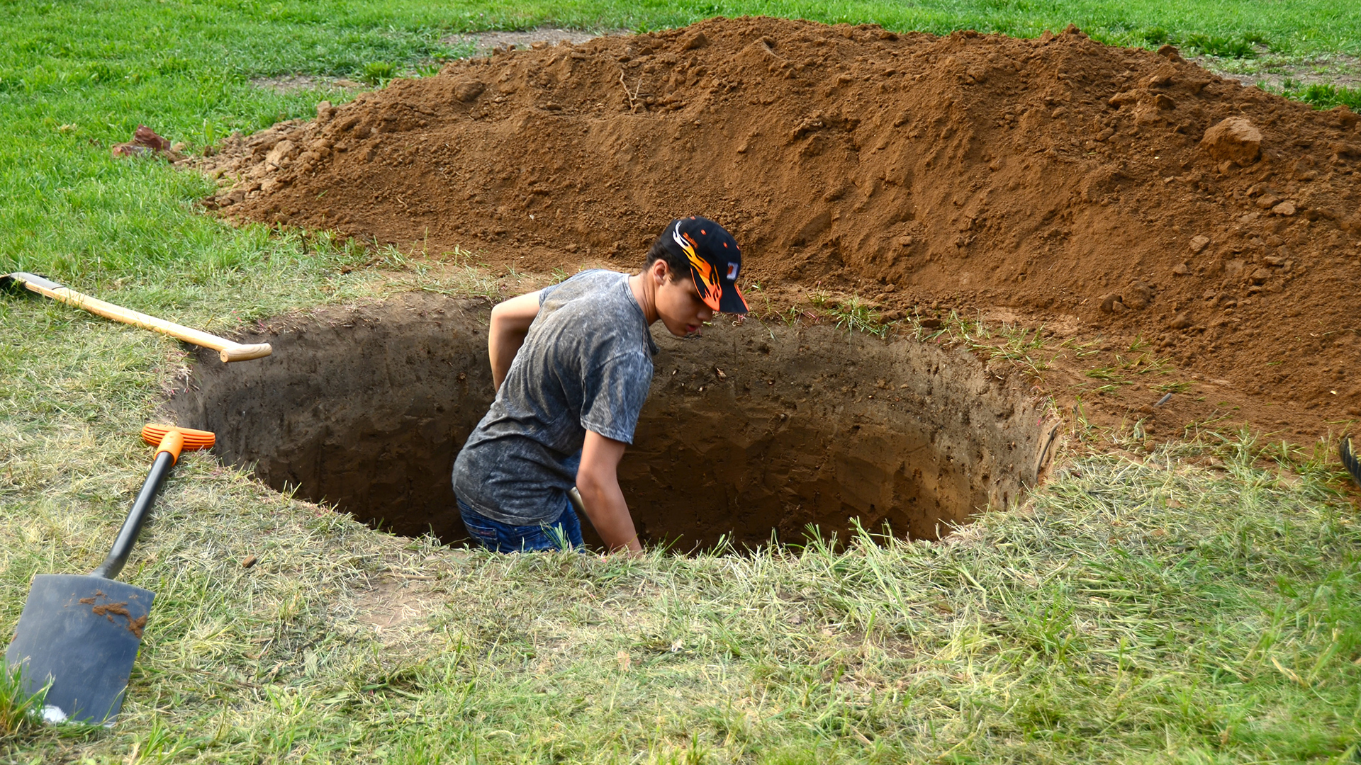 A colour photograph of a person digging a hole. The person is standing in a waist-deep hole, with mounds of dirt around them.