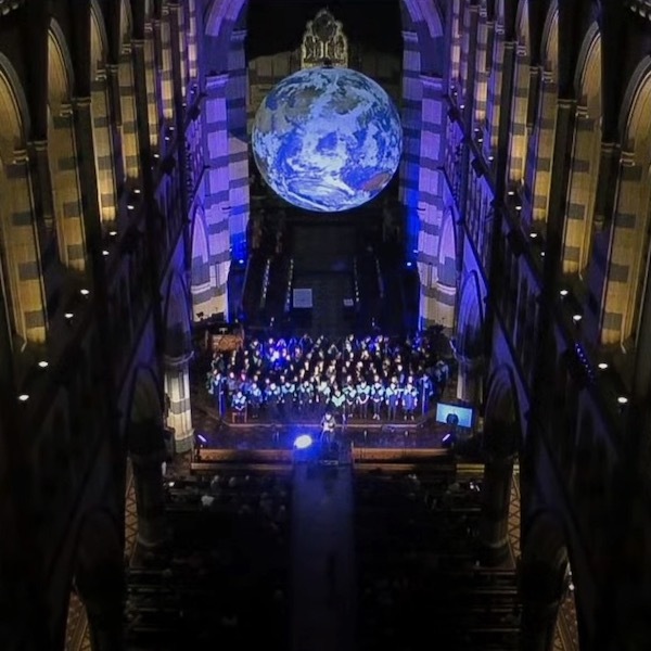 The interior of a gothic cathedral looking from above. Below is a choir singing below a giant revolving inflatable Earth.