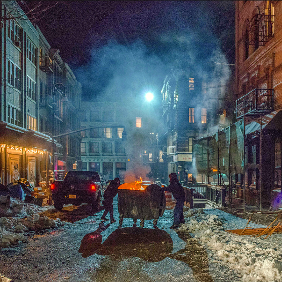 Two silhouetted people dressed in hoodies and jeans push an open dumpster filled with fire down an empty street at night. Snow is lumped on the roadside, amongst full bin bags and discarded objects.
