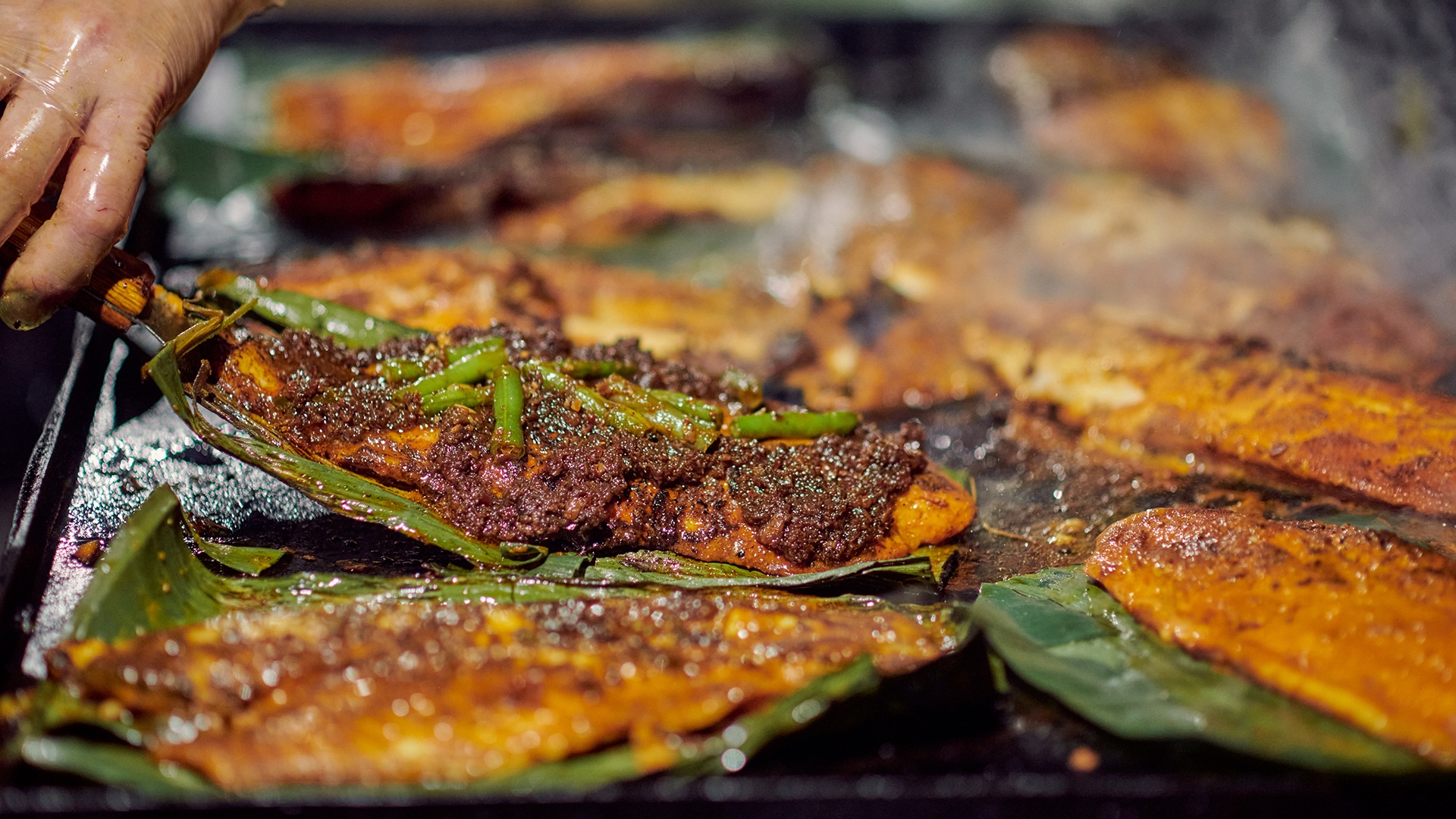 Sautead skewers on banana leaves being cooked on a hot grill. A hand repositions one of the skewers. 